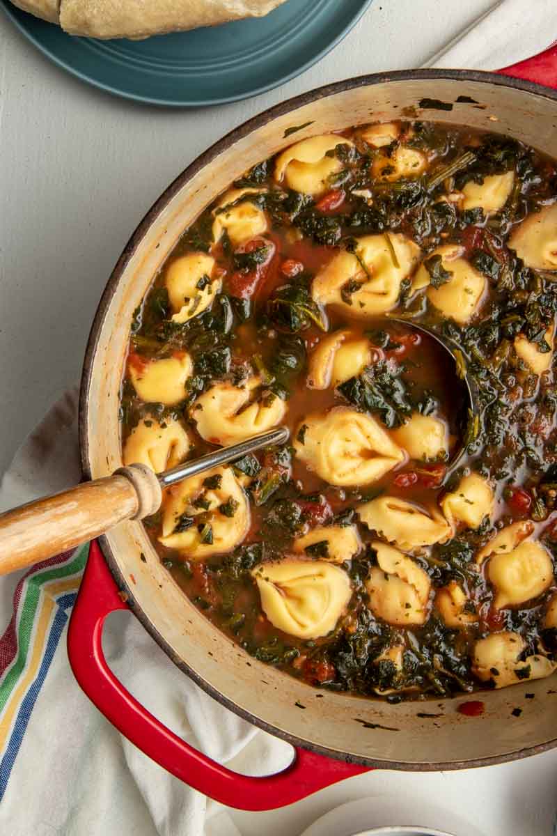 Top view of an enameled dutch oven with red handles filled with spinach tortellini soup and a serving ladle.