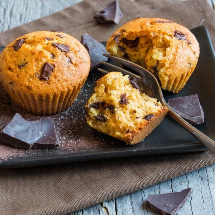 A fork splits one of two muffins on a black plate with cocoa powder and pieces of chocolate around them.