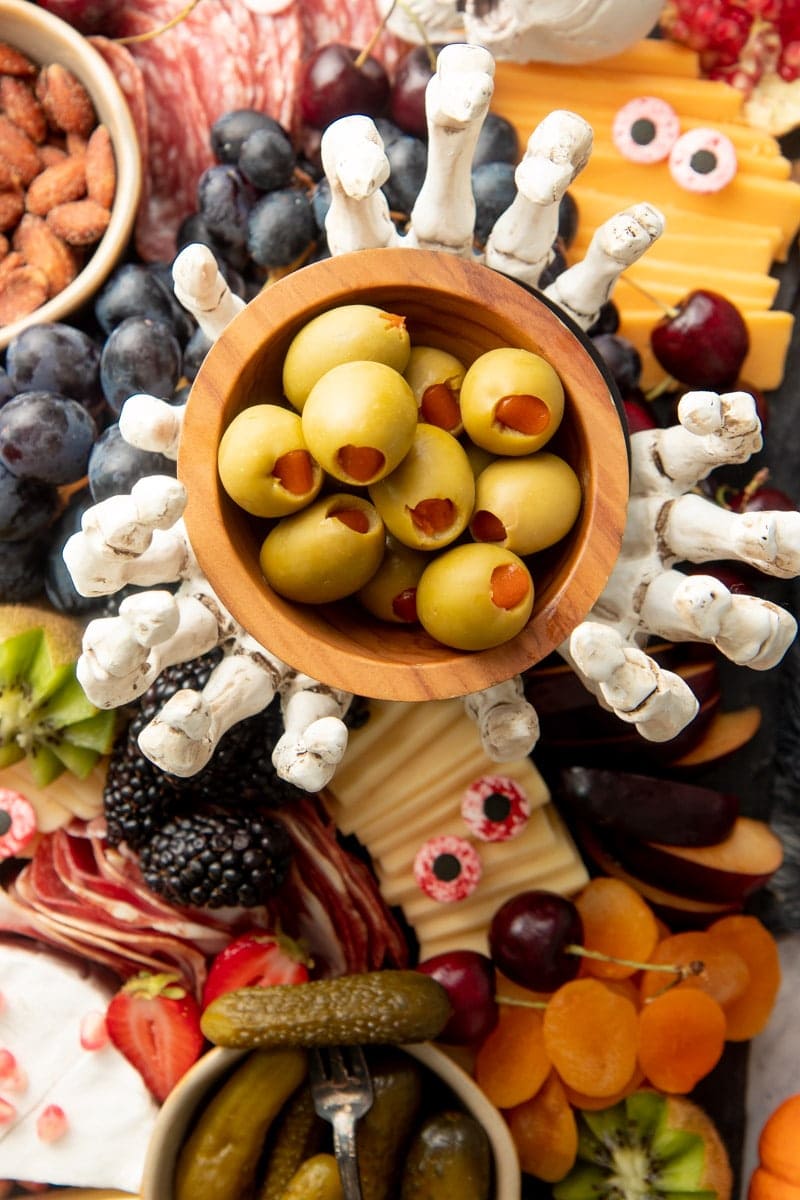 Three skeleton hands hold up a bowl of stuffed olives on a cheese board filled with meats, cheeses, fruits, nuts, and crackers.