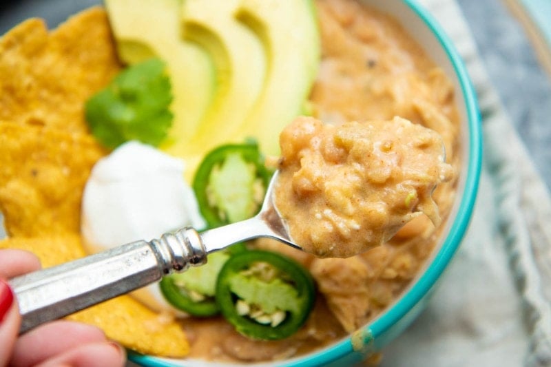 Close view of a spoonful of white chicken chili above a bowl garnished with avocado and jalapeno slices.