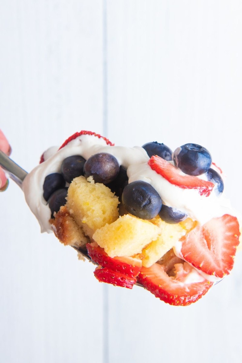 A spoon loaded with cubes of cake, slices of strawberries, fresh blueberries, and cream cheese whipped cream.