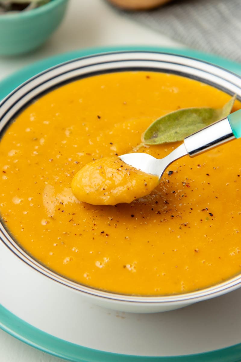 A spoonful of roasted butternut squash soup is lifted out of a full bowl garnished with fresh cracked black pepper and sage.