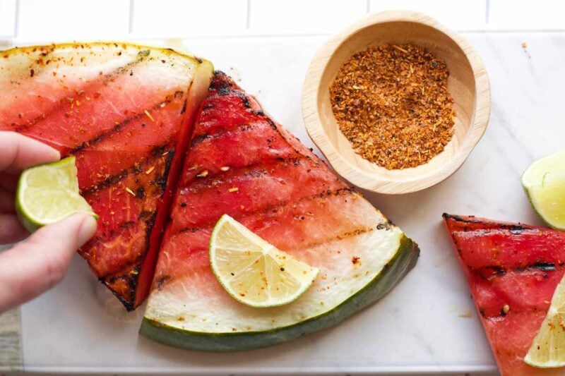 A hand squeezes a slice of lime over two wedges of melon, a ramekin of tajin seasoning nearby.