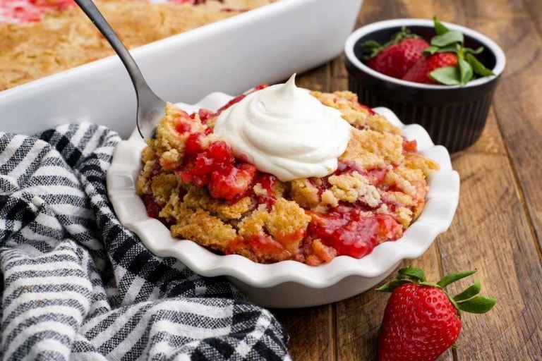 Close view of a fork scooping a bite of strawberry dump cake from a white bowl.