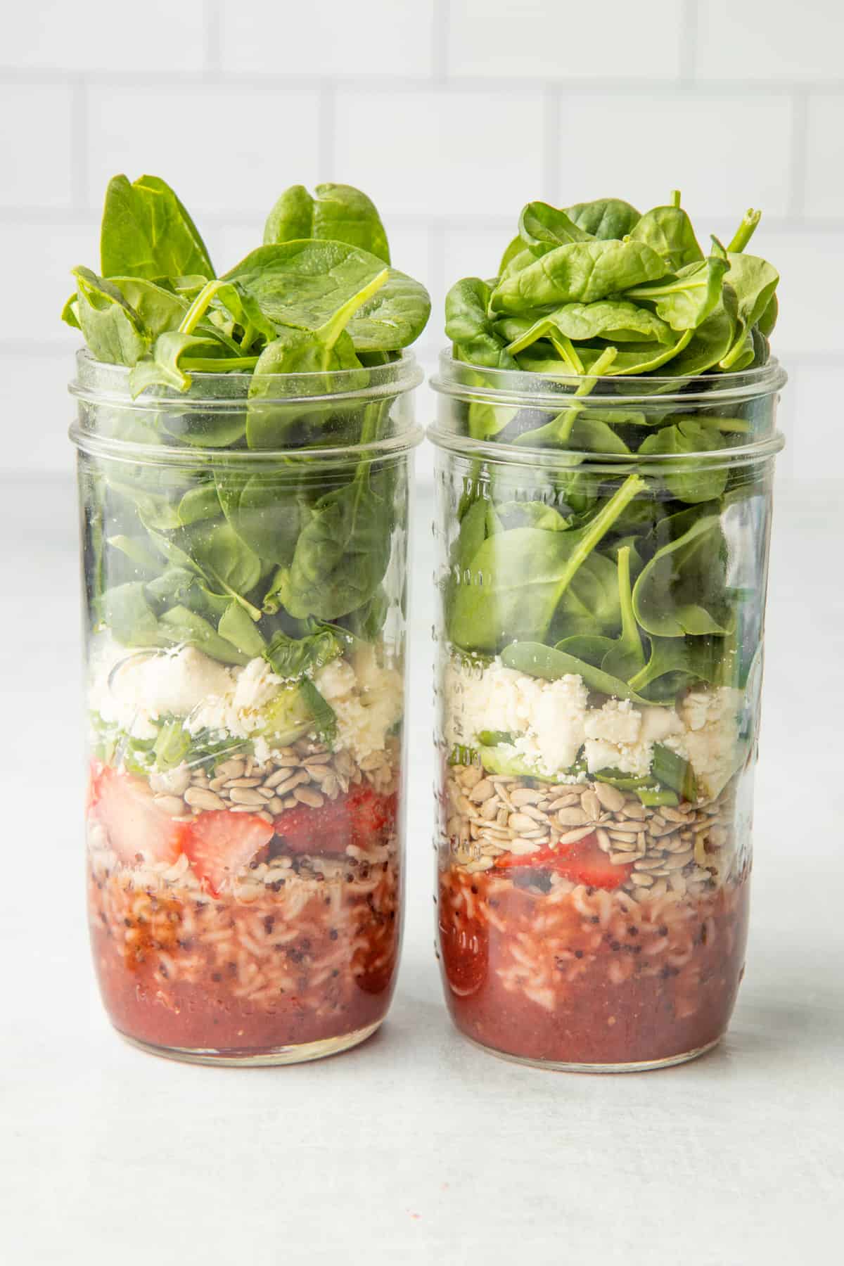 Two mason jars sit side-by-side, layered with components of strawberry spinach salads.