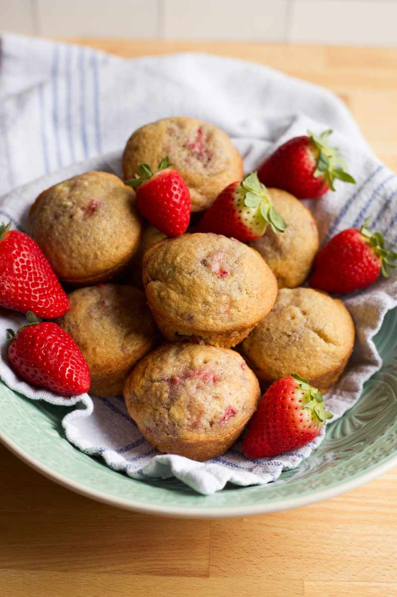 A tea towel-lined bowl is filled with strawberry muffins and fresh berries.