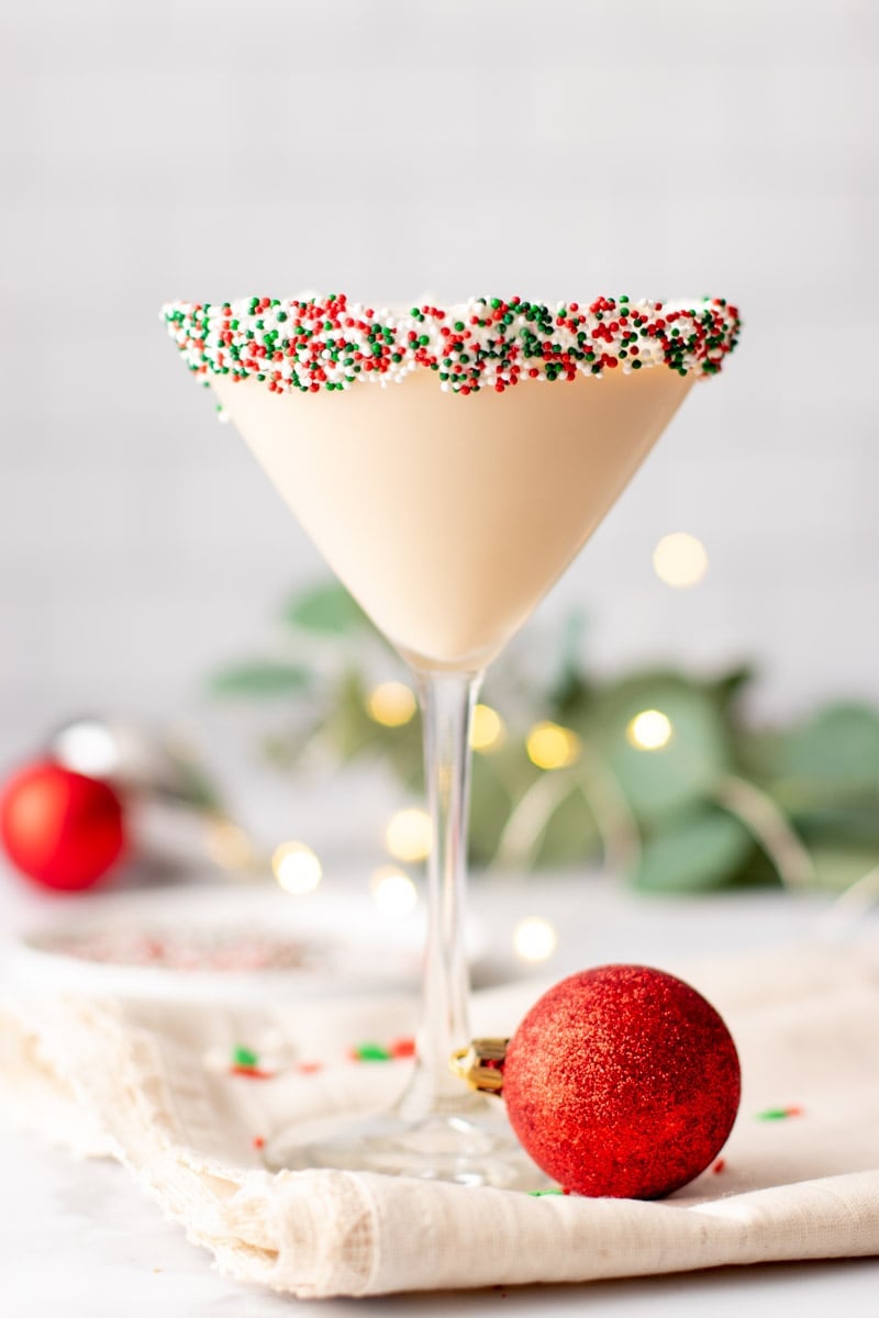 A Christmas martini with a sprinkle rim stands with a red Christmas ball at its foot.