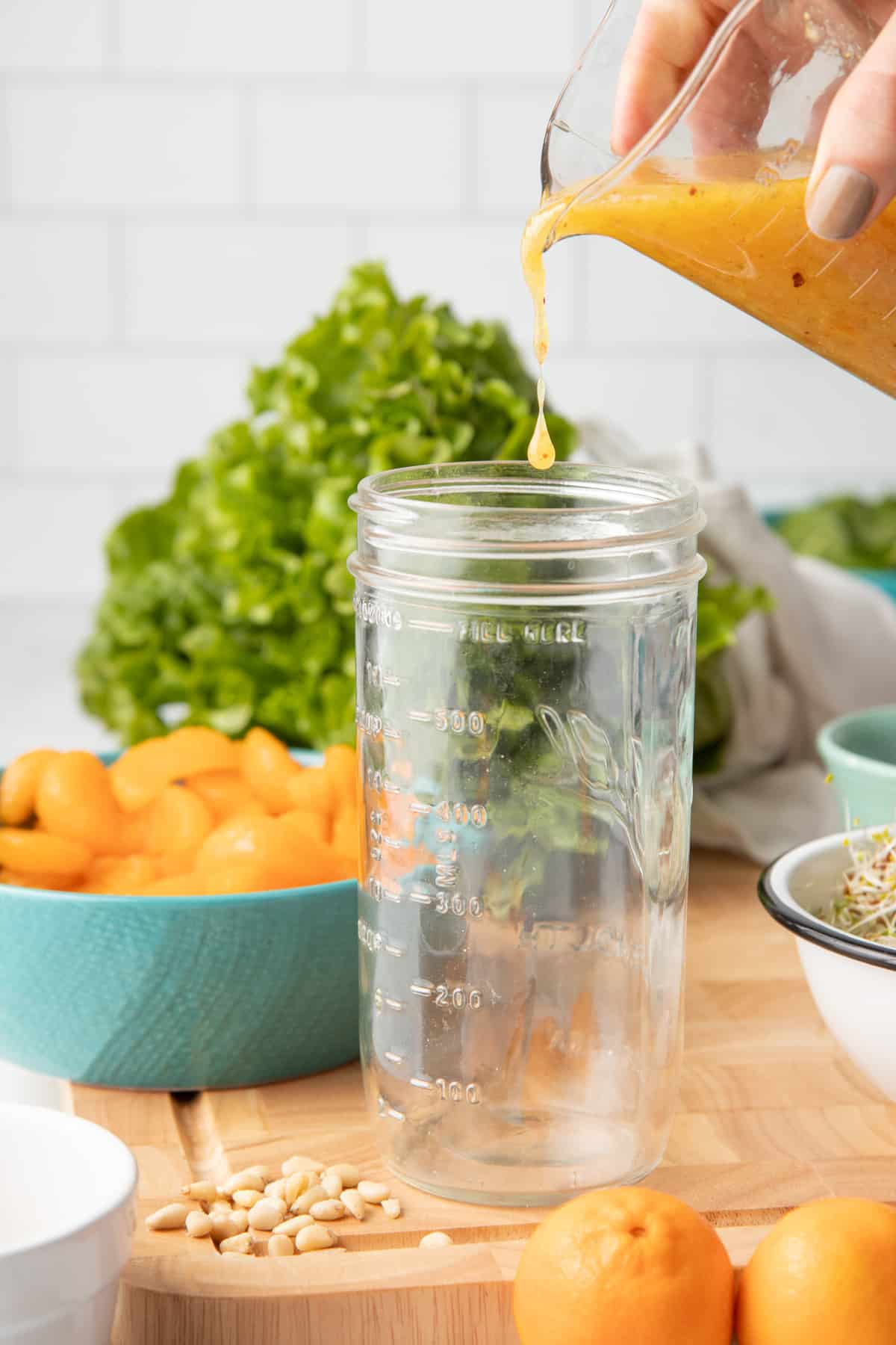 A hand pours orange marmalade vinaigrette from a measuring cup into the bottom of a mason jar.