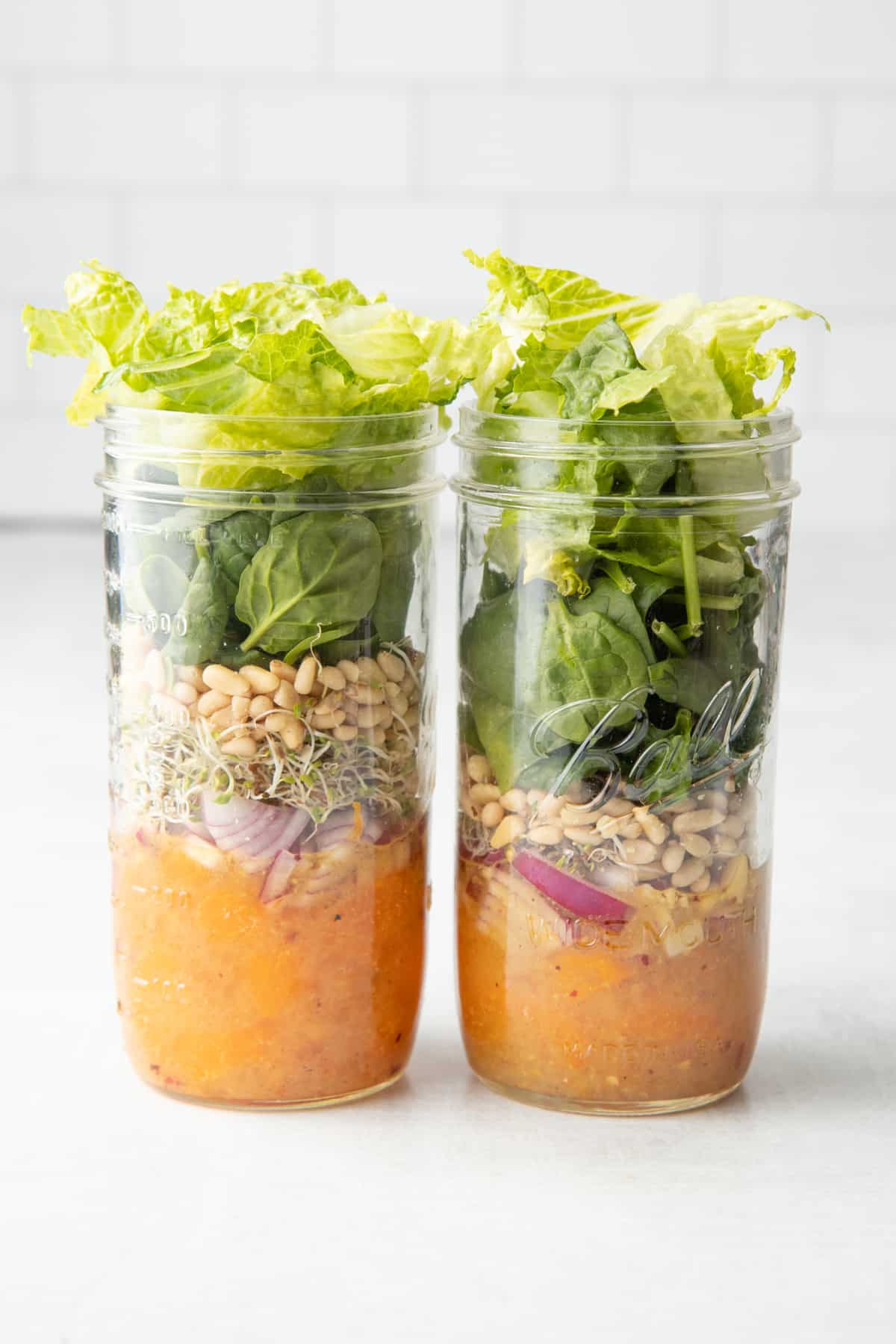 Two jars full of salad components for a citrus salad - dressing, mandarin slices, onions, sprouts, pine nuts, and greens.