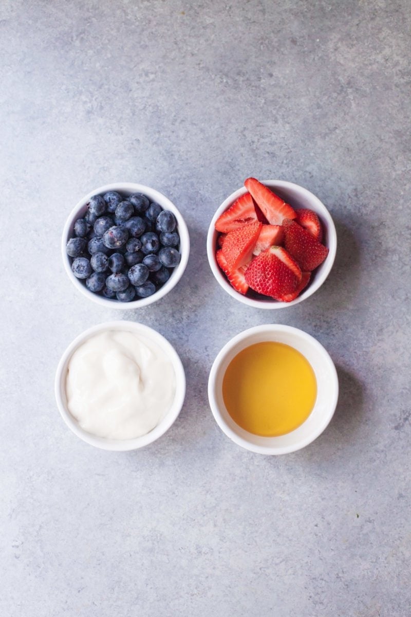 Ingredients in individual white bowls - blueberries, strawberries, yogurt, and honey