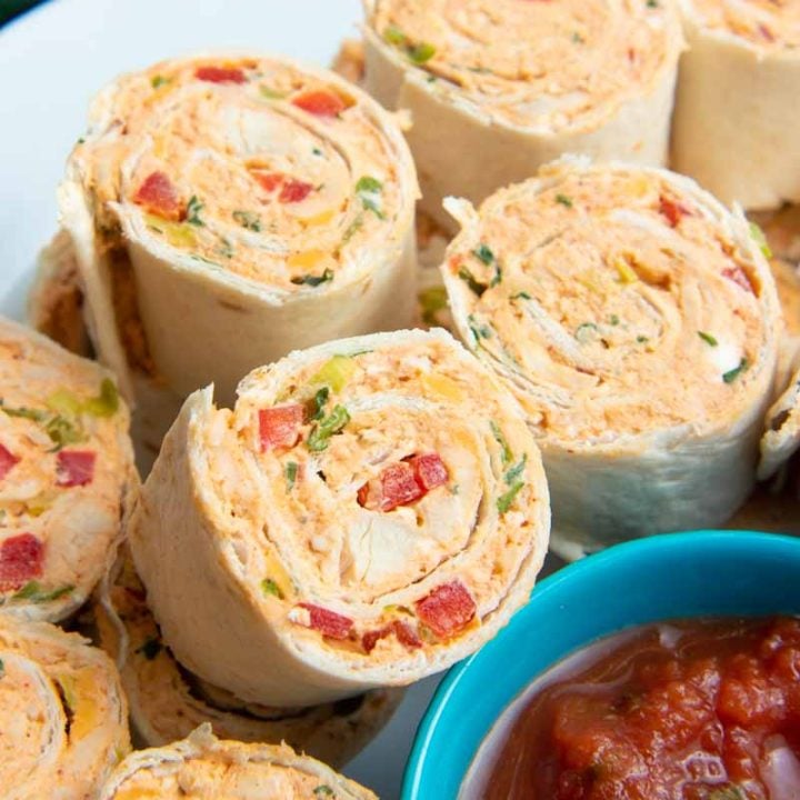 Close view of taco pinwheels piled on a white plate around a small teal bowl of salsa for dipping.