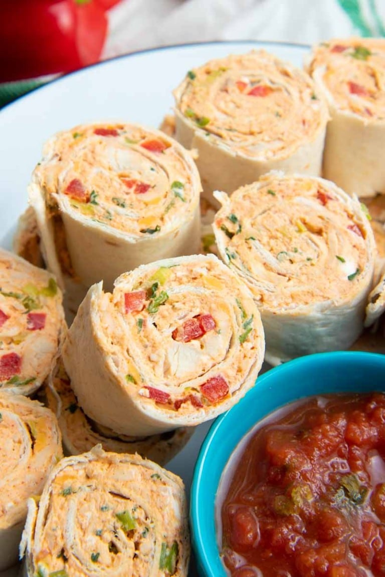 Close view of taco pinwheels piled on a white plate around a small teal bowl of salsa for dipping.