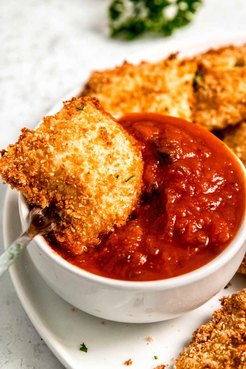 A fork dips a toasted ravioli into a bowl of marinara sauce.