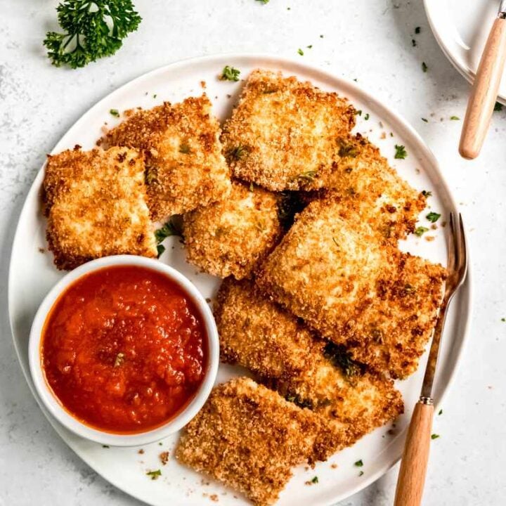 Overhead of toasted ravioli served on a white plate with a small bowl of marinara sauce and serving forks nearby.