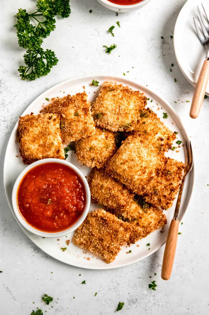 Overhead of toasted ravioli served on a white plate with a small bowl of marinara sauce and serving forks nearby.