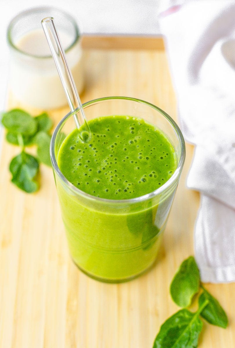 Close view of spinach smoothie in a glass with glass straw surrounded by fresh spinach leaves.