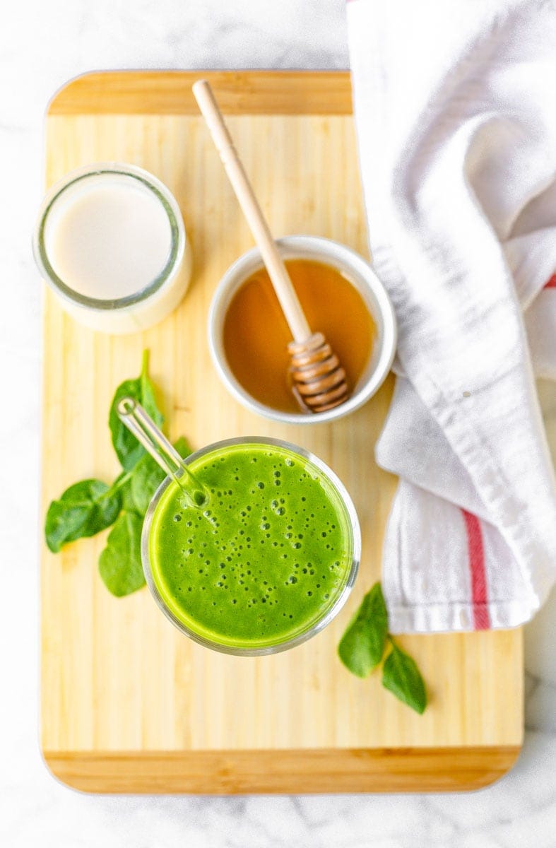 Overhead of spinach smoothie on a cutting board with honey, fresh spinach leaves, and milk.