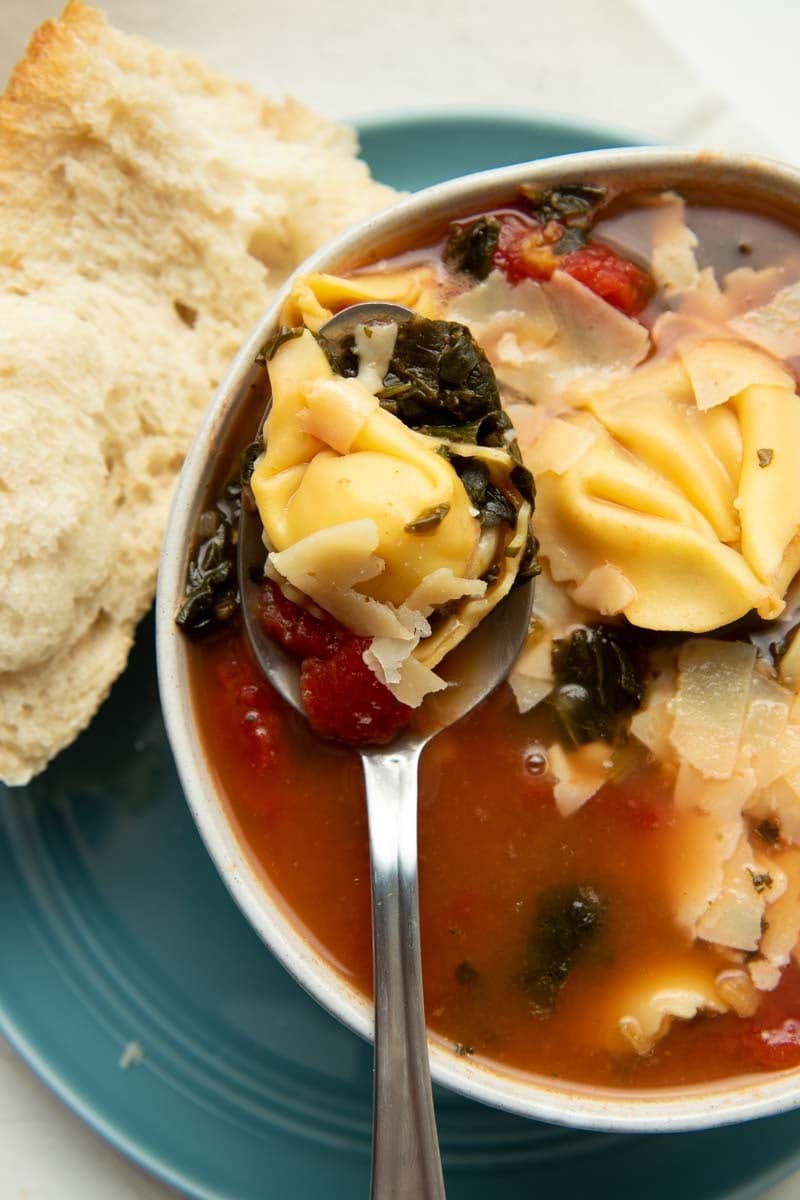 A spoon with a full bite of soup rests across a serving bowl of the brothy soup, a hunk of bread alongside.