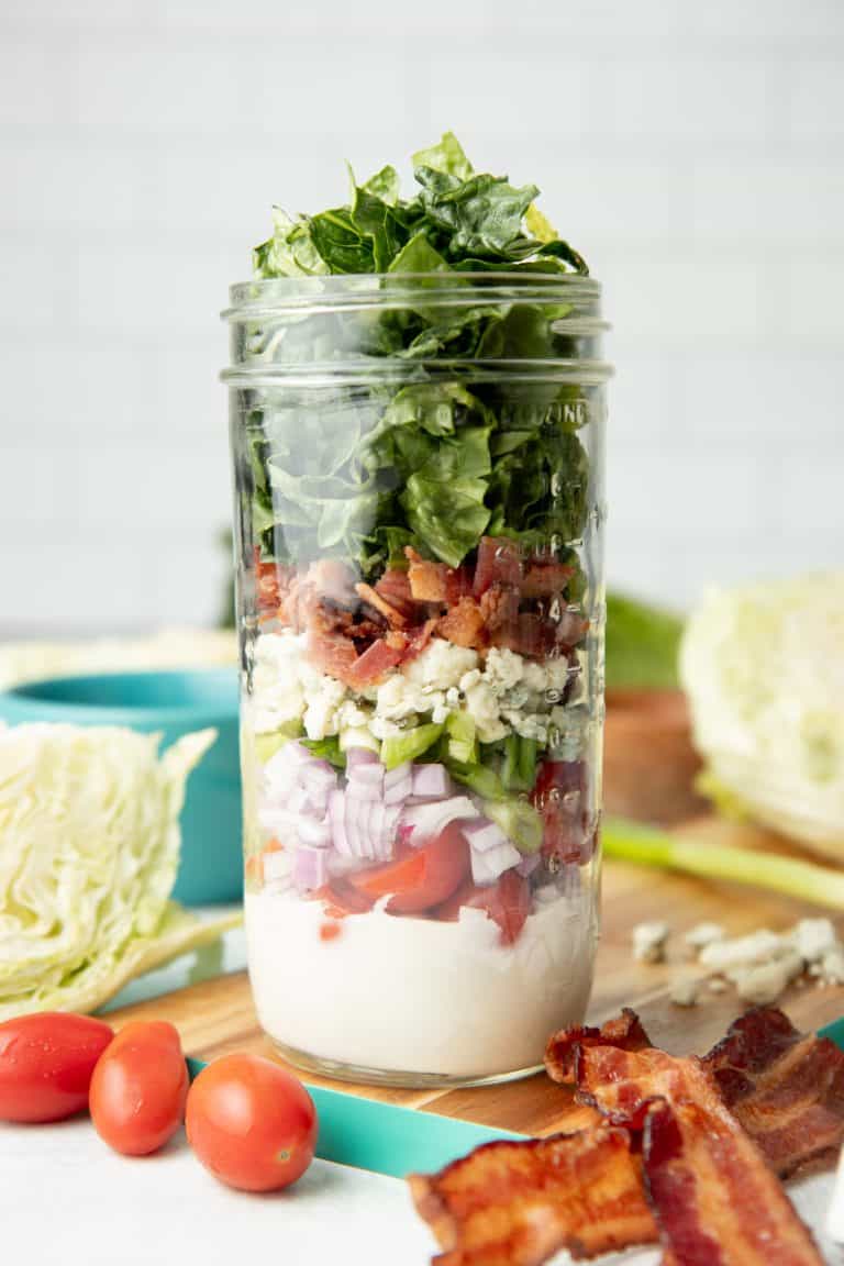 Components of a wedge salad are layered in a glass mason jar, which sits on a teal-edged cutting board. Additional ingredients surround the jar.