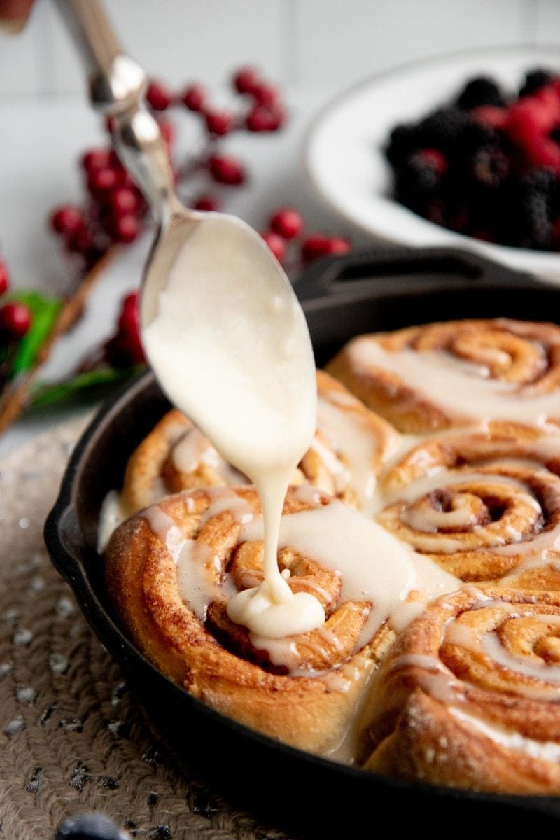 Spoon drizzling icing onto baked vegan cinnamon rolls in a cast iron skillet.