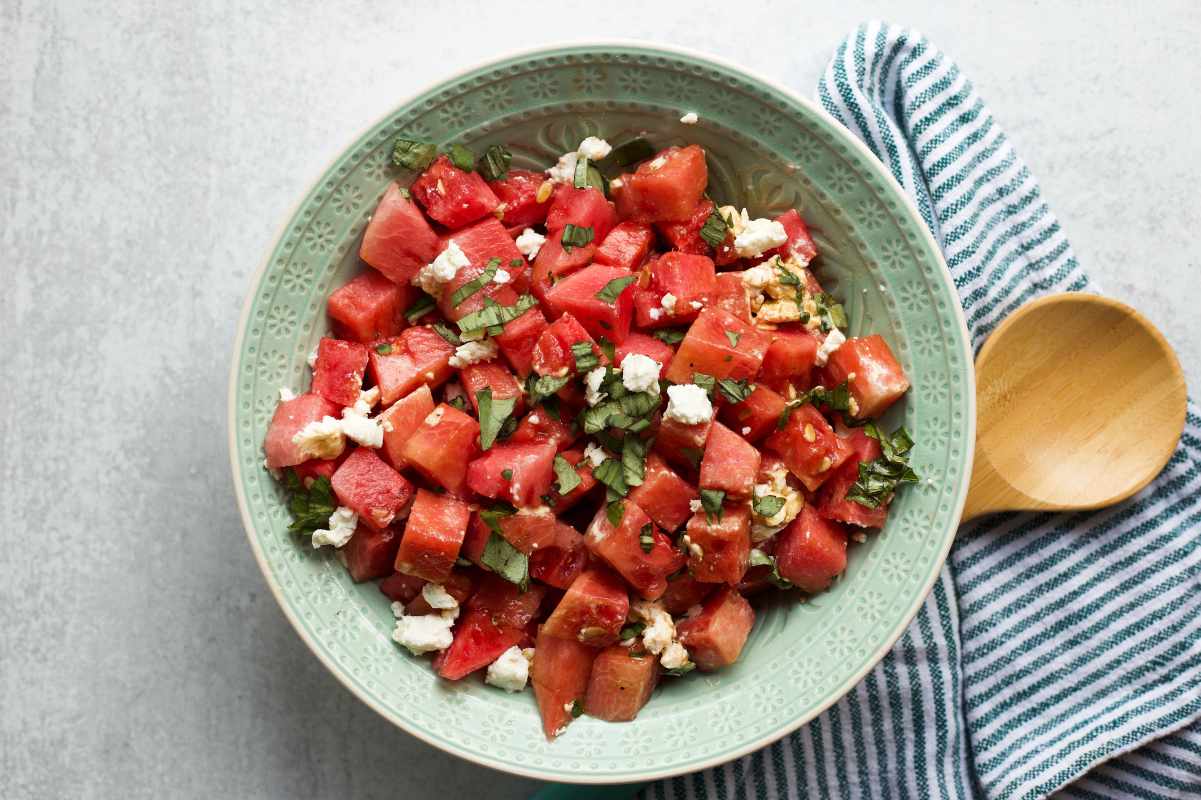 A wooden spoon rests on a striped linen napkin beside a decorative bowl filled with watermelon basil salad.