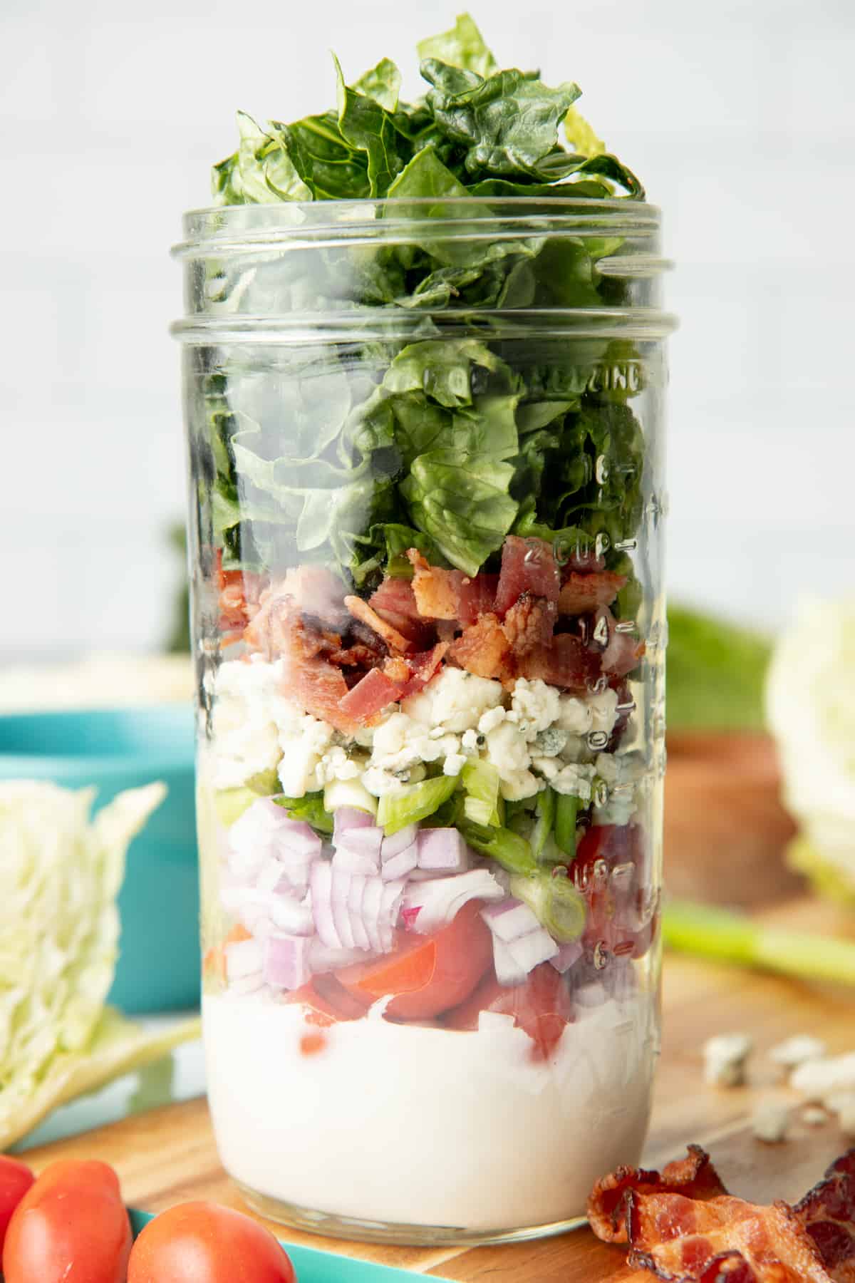 Components of a wedge salad are layered in a glass mason jar, which sits among extra ingredients.