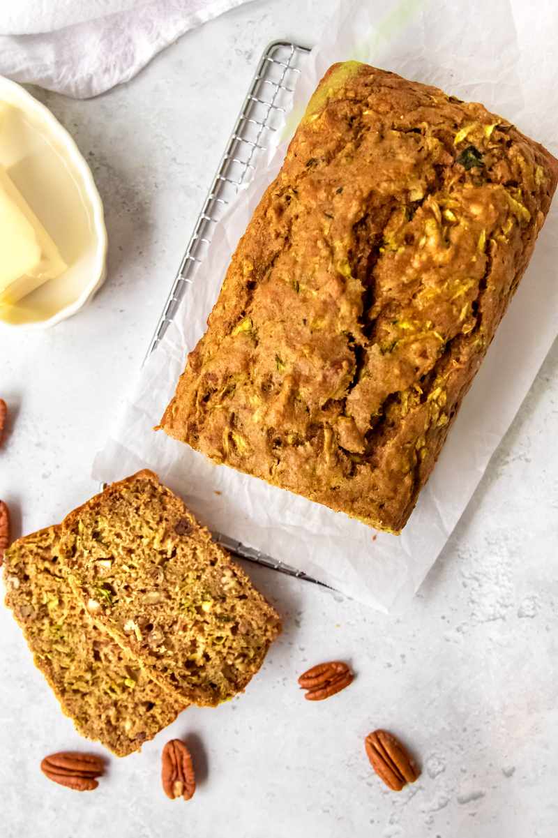 Top view of a loaf of whole wheat zucchini bread with two slices cut, butter and pecan halves nearby.