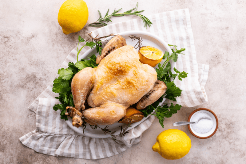 Roast chicken on a white platter with lemons, parsley, rosemary, and salt