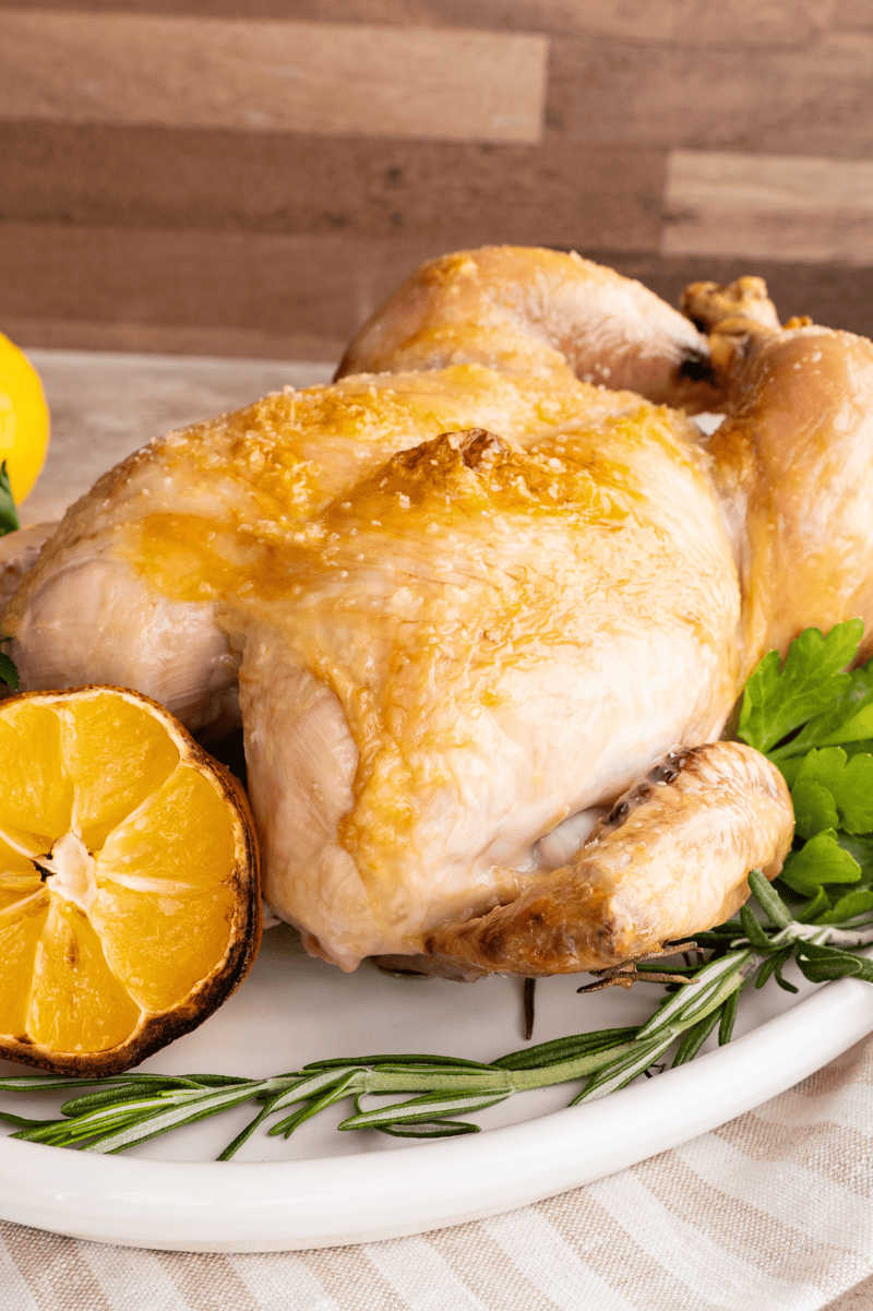 Cooked poultry with browned, crispy skin on a white platter with fresh herbs and a halved lemon